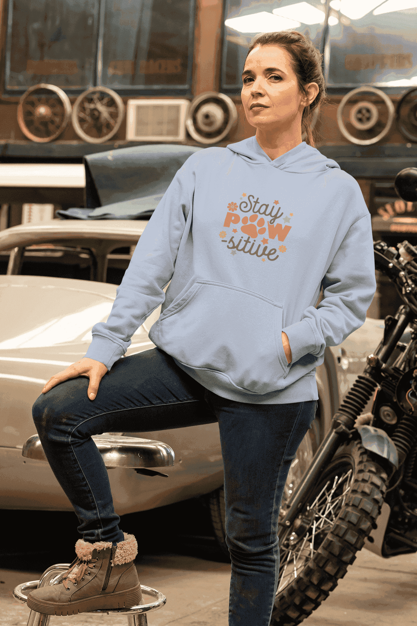 Woman wearing light blue Stay Pawsitive Unisex Hoodie with paw graphic leaning on vintage car in garage