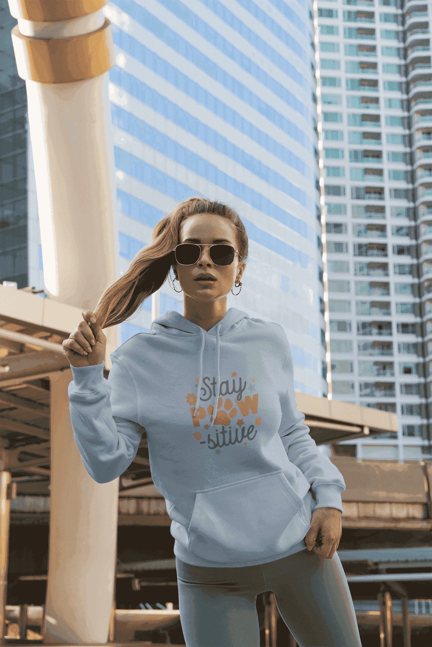 Young woman wearing Stay Pawsitive Unisex Hoodie light gray with paw print design and sunglasses in city