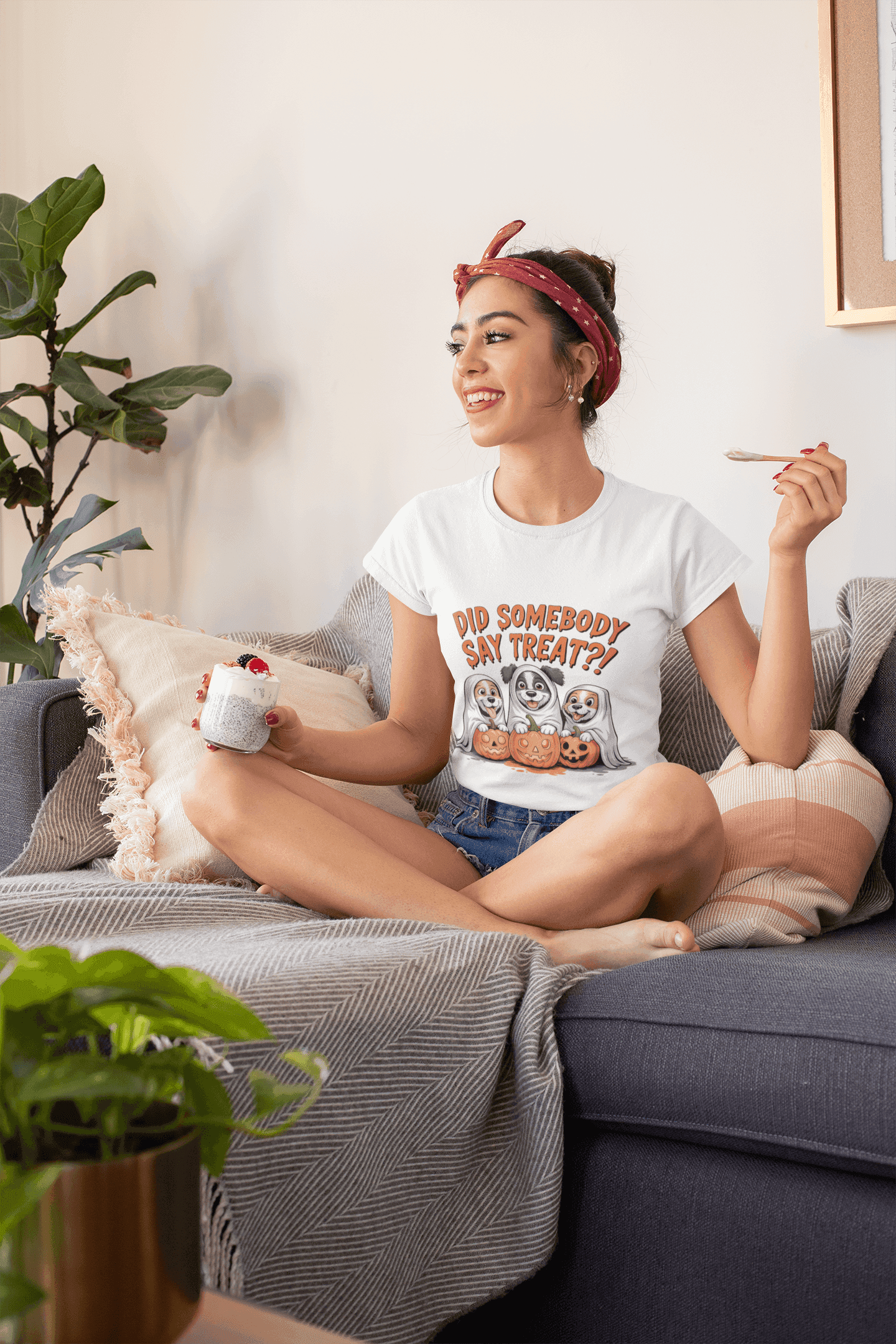 Smiling woman on sofa wearing Printify Spooky Halloween Dog Tee white shirt with three dogs and treats graphic holding dessert