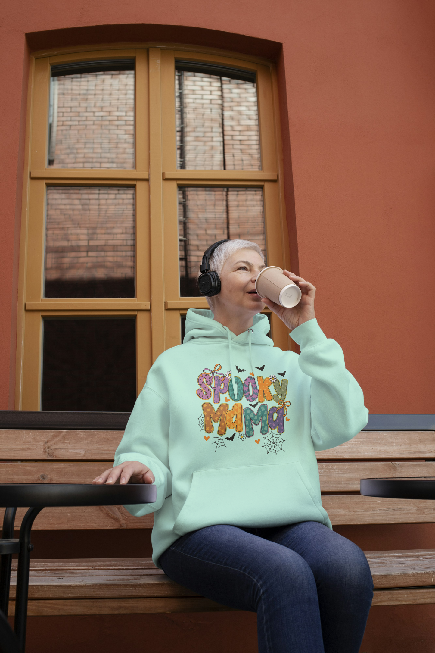 Person wearing a 'Spooky Mama' hoodie sitting on a bench outdoors.