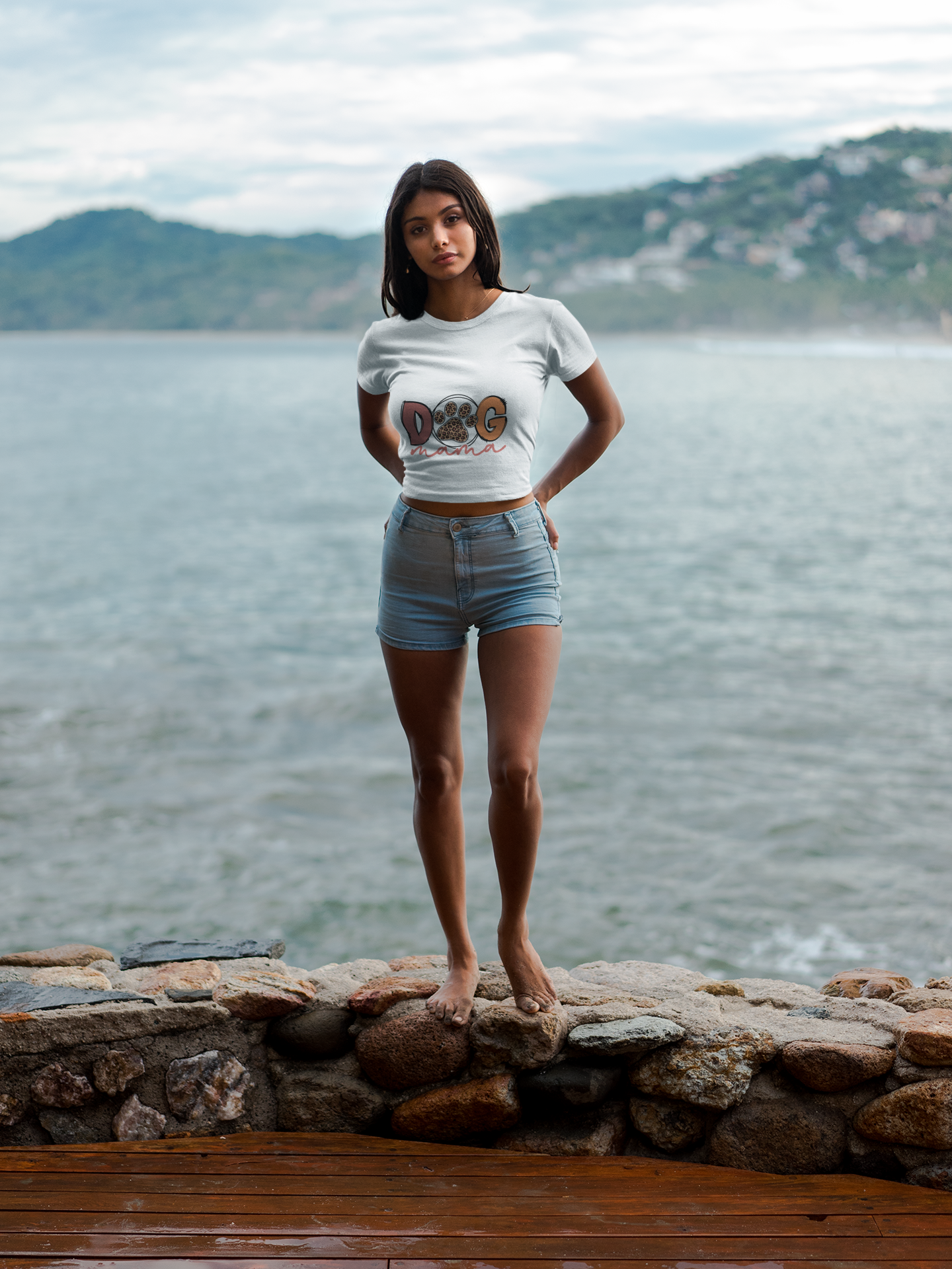 Woman standing barefoot by the sea wearing a fitted Printify Dog Mama Tee and denim shorts