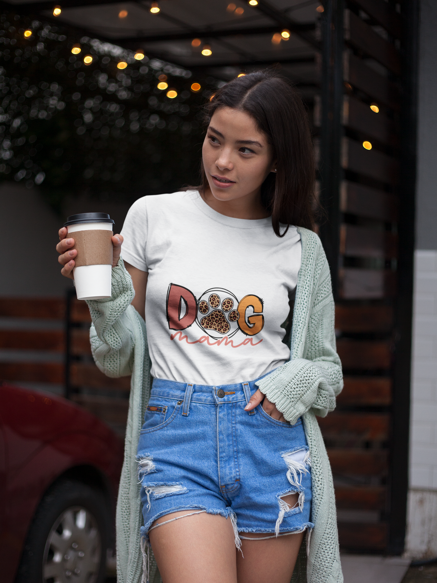 Young woman wearing white Dog Mama Tee with paw print design, green cardigan, ripped denim shorts, holding coffee