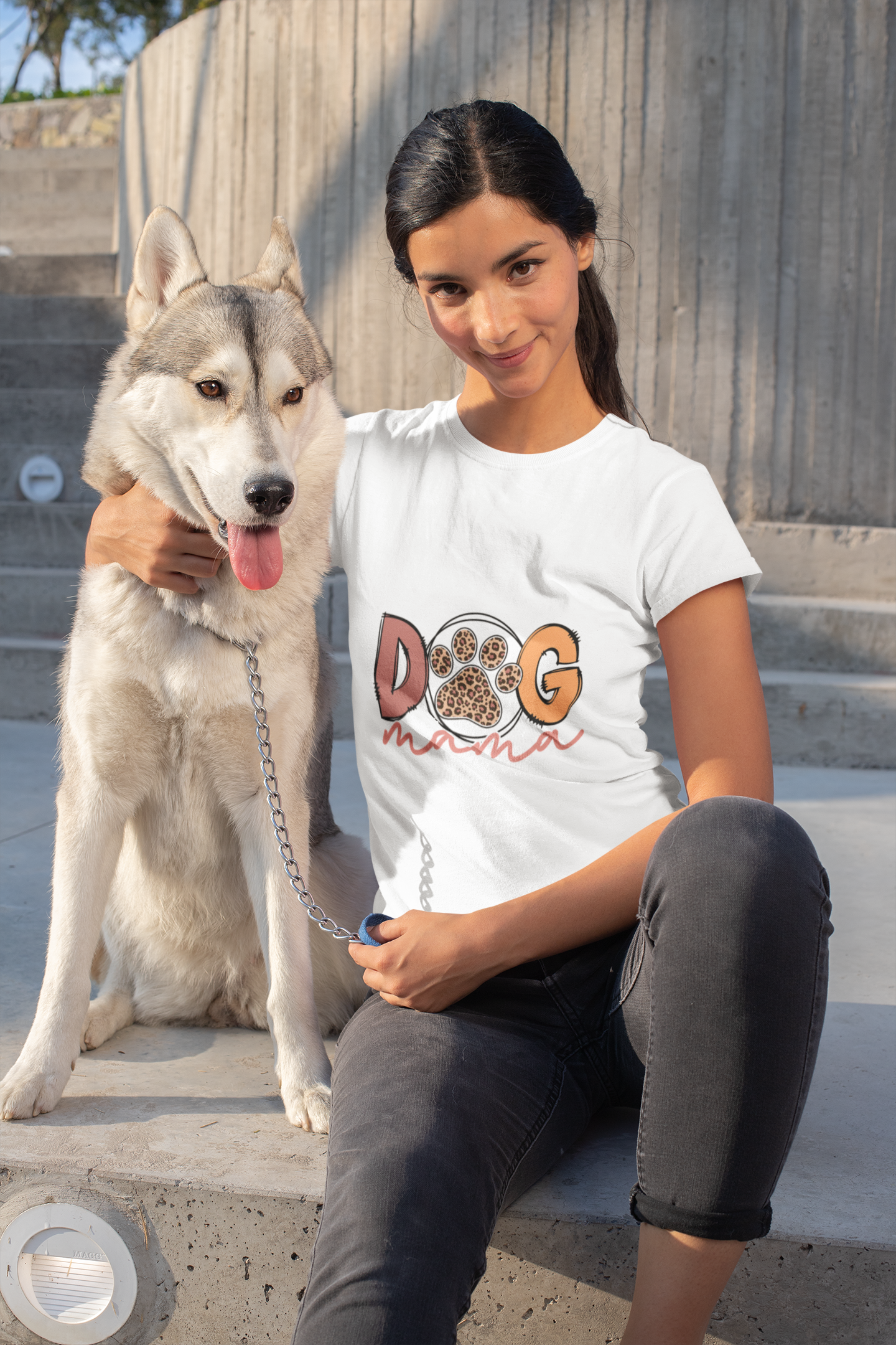Smiling woman wearing a white Printify Dog Mama Tee sitting on concrete steps with a husky