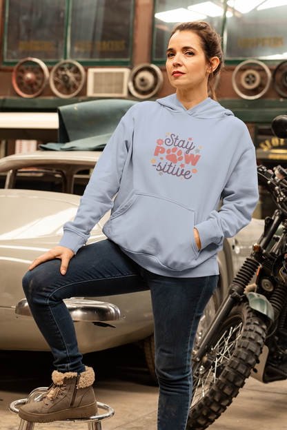 Woman wearing light blue Stay Pawsitive Unisex Hoodie with paw graphic leaning on vintage car in garage