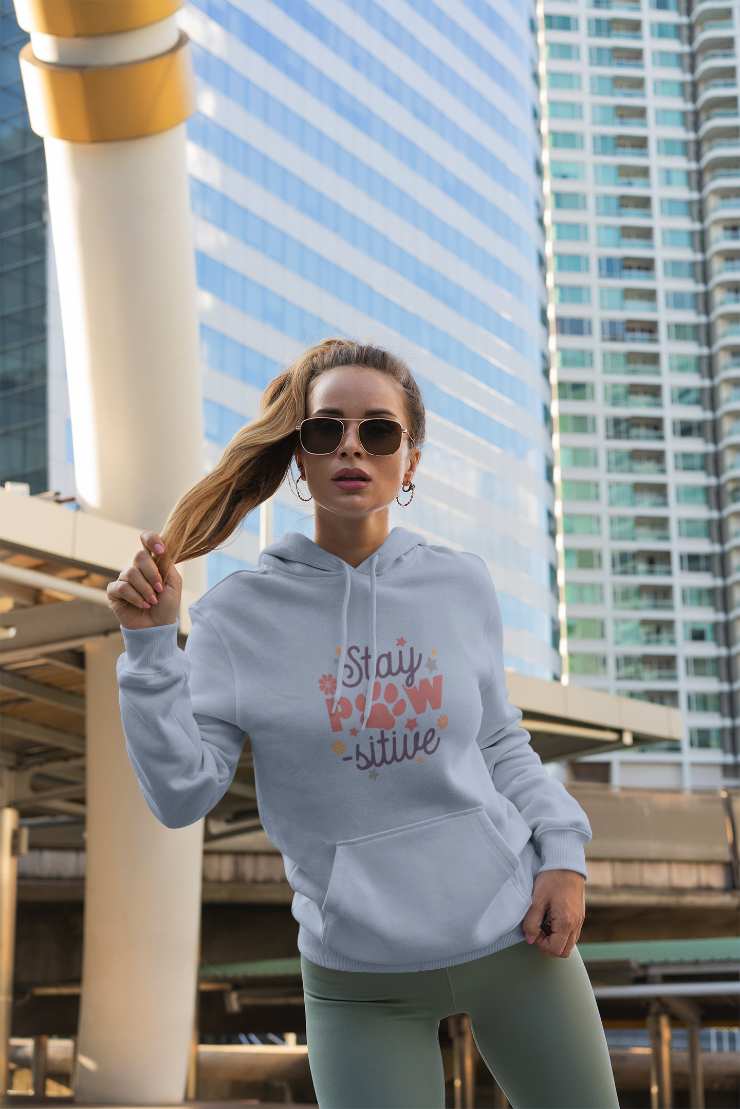 Young woman wearing Stay Pawsitive Unisex Hoodie light gray with paw print design and sunglasses in city