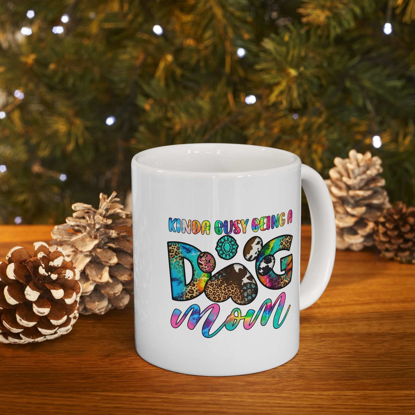 White ceramic Pet Owner Coffee Cup with colorful Kinda Busy Being a Dog Mom design and paw print on wooden table with pine cones