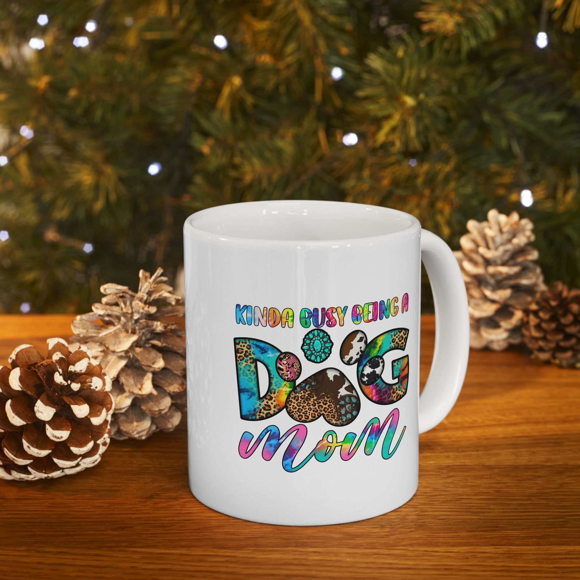 White ceramic Pet Owner Coffee Cup with colorful Kinda Busy Being a Dog Mom design and paw print on wooden table with pine cones