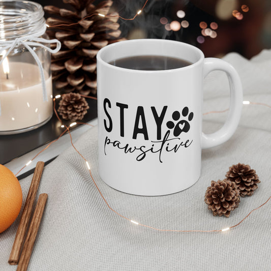 White Stay Pawsitive Mug with black text and paw print filled with coffee surrounded by pinecones and candle