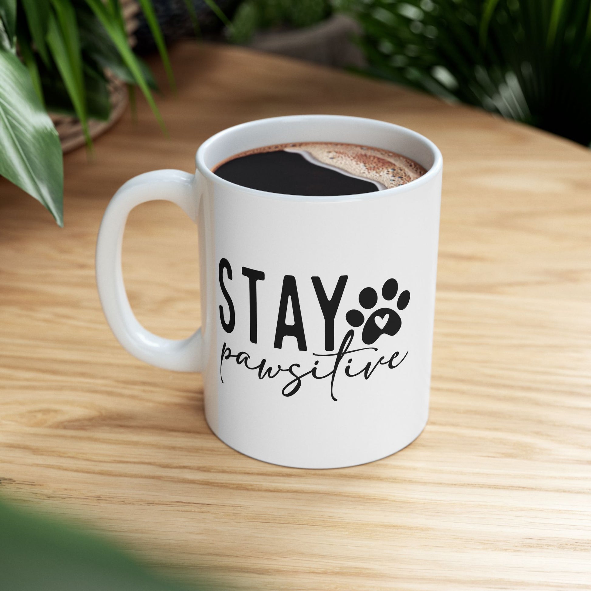 White ceramic Stay Pawsitive Mug with black text and paw print design on wooden table